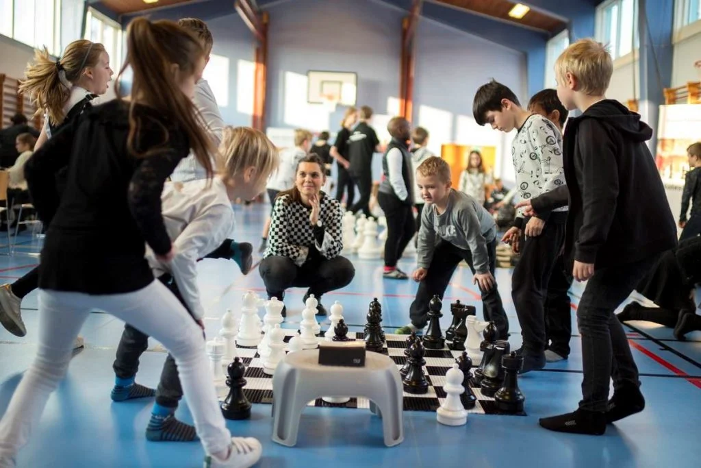 Elever och en lärare samlas runt ett stort schackbräde i en gymnastiksal.