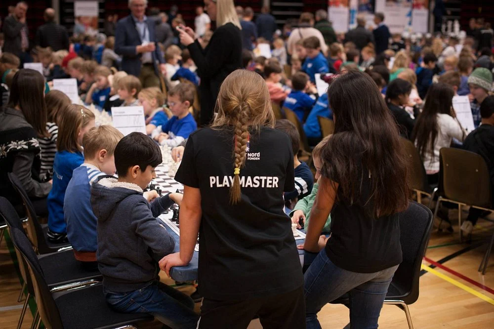 Unga PLAYMASTERS hjälper elever vid schackbräden under ett gemensamt evenemang.